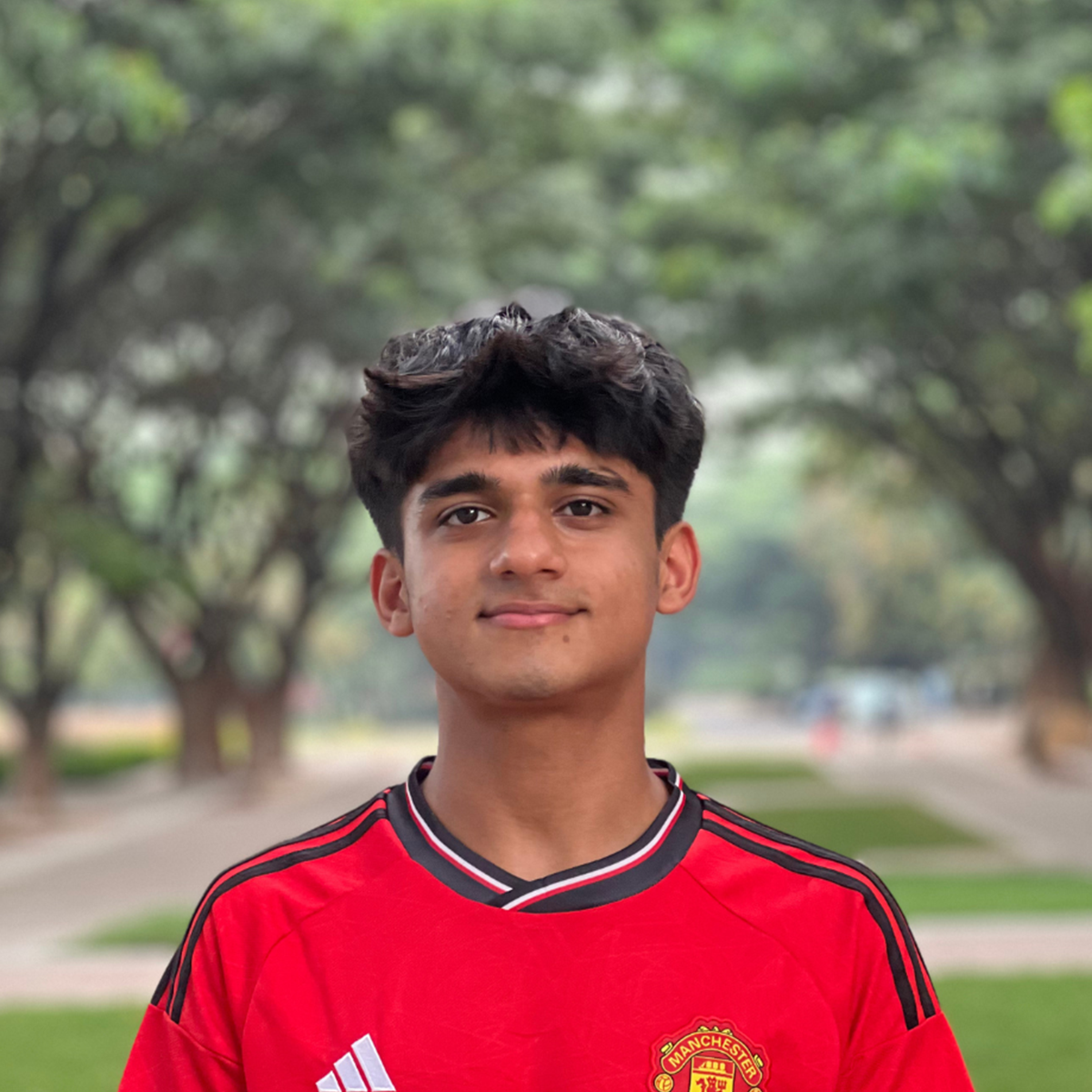 Outdoor portrait of a young man in a red football shirt against a leafy background.